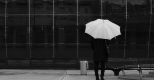 Eine einsame Person mit einem Regenschirm steht vor einer modernen Glaswand und regt zur Introspektion an.