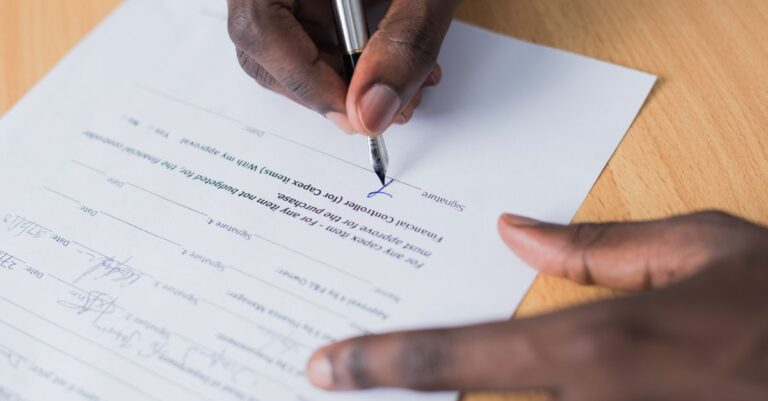 Eine Nahaufnahme der Hand einer Person, die einen Geschäftsvertrag auf einem Tisch mit einem Stift unterschreibt.